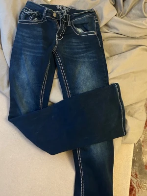 Mörkblå jeans med kontrastsömmar - Snygga mörkblå jeans från Infinity med vita kontrastsömmar och dekorativa bakfickor med lock och knapp. Jeansen har klassisk femficksmodell och lätt slitningar framtill för en cool look. Tillverkade i denim med stretch för skön passform.