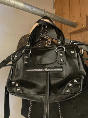 Svart handväska med nitar och kedjedetaljer - Cool svart handväska i glansigt material med silvriga nitar och kedjedetaljer. Väskan har dubbla handtag, dragkedja framtill och dekorativa metalldetaljer. Perfekt för dig som gillar en edgy och trendig look. Inte min stil 