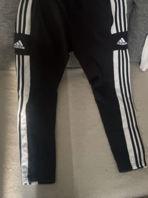 Svarta Adidas träningsbyxor - Säljer ett par svarta träningsbyxor från Adidas med klassiska vita ränder längs sidorna och Adidas-logga på båda benen. Byxorna har smal passform och är perfekta för träning eller sport. Materialet är polyester för bra rörelsefrihet.