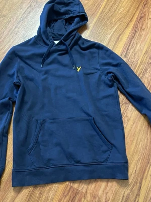 Mörkblå hoodie från Lyle & Scott - Snygg mörkblå hoodie från Lyle & Scott med klassisk gul logga på bröstet. Hoodien har dragsko i huvan, känguruficka framtill och ribbade muddar. Tillverkad i mjukt bomullsmaterial som är skönt att ha på sig.