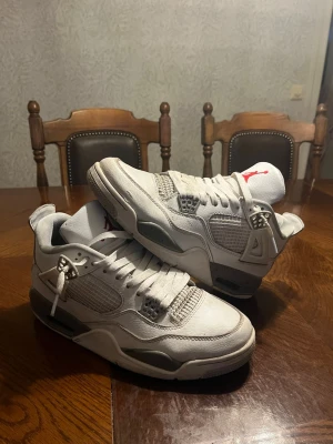 Air Jordan 4 Retro White Oreo - Snygga Air Jordan 4 Retro White Oreo sneakers i vitt läder med grå och svarta detaljer. Klassisk siluett med meshpaneler, plastvingar och synlig Air-sula. Röd Jumpman-logga på plösen och stänkmönster på mellansulan. Perfekta för dig som gillar streetwear och ikoniska sneakers.