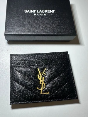 Svart Saint Laurent kortfodral - Snyggt svart kortfodral från Saint Laurent i präglat läder med guldigt YSL-logo framtill. Fodralet har flera kortfack och en stilren, lyxig design som passar perfekt i fickan. Kommer med originalask.