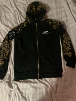 Svart hoodie från Icono Couture - Svart hoodie med dragkedja från Icono Couture. Snygga guldmönster på ärmar och huva, samt vit logga på bröstet. Hoodien har fickor framtill och justerbar huva med snören. Perfekt för dig som gillar streetstyle och unika detaljer.