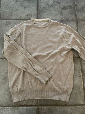 Beige rundhalsad Stenströms tröja - Beige stickad tröja med rund halsringning och ribbade muddar vid ärmslut och nederkant. Tröjan har förstärkta armbågslappar i samma färg för en stilren look. Perfekt för dig som gillar enkel och clean stil.