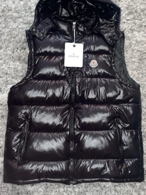 Svart dunväst från Moncler - Snygg svart dunväst från Moncler med glansig finish och klassisk logotyp på bröstet. Västen har dragkedja framtill, två fickor med dragkedja och hög krage. Perfekt för lager-på-lager och streetstyle.