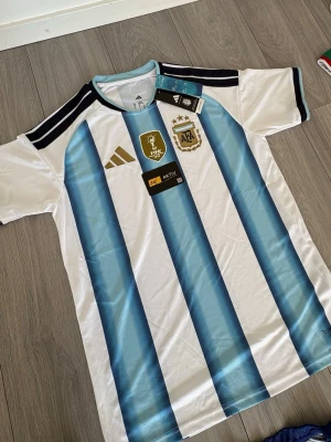 Argentina matchtröja Adidas blåvit - Säljer en officiell Argentina matchtröja från Adidas med klassiska blå och vita ränder, guldfärgade detaljer och FIFA-märke på bröstet. Tröjan har rund hals, korta ärmar och är tillverkad i ett lätt funktionsmaterial. Perfekt för fotbollsfans!