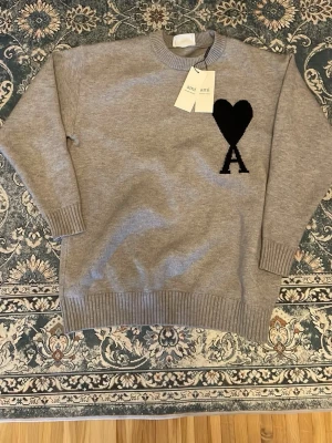 Grå stickad tröja från Ami Paris - Snygg grå stickad tröja från Ami Paris med svart hjärta och bokstaven A på bröstet. Tröjan har rund halsringning, ribbade muddar och är tillverkad i mjukt material. Perfekt för en stilren och trendig look.