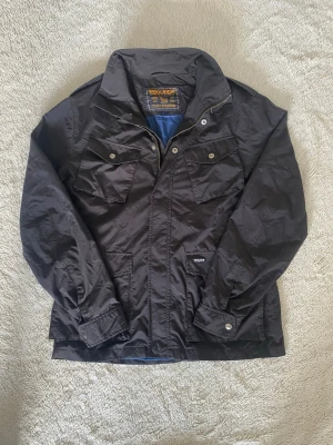 Svart vindjacka från Woolrich - Svart vindjacka från Woolrich med klassisk krage, fyra fickor framtill och tryckknappar. Jackan har dragkedja och är tillverkad i ett lätt, vattenavvisande material. Perfekt för blåsiga dagar och enkel att matcha med olika outfits.