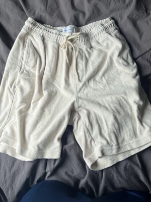 Beiga frottéshorts med snörning - Snygga beiga shorts i mjuk frotté med elastisk midja och snörning framtill. Shortsen har sidofickor och en bakficka, perfekt för en avslappnad stil under varma dagar.