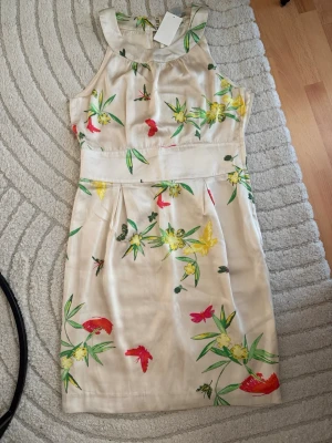 Blommig ljus kortklänning - Snygg ljus kortklänning med blommigt mönster i grönt, gult och rött. Klänningen är ärmlös och har en rak, enkel passform med markerad midja. Perfekt för varma dagar och somriga tillfällen.