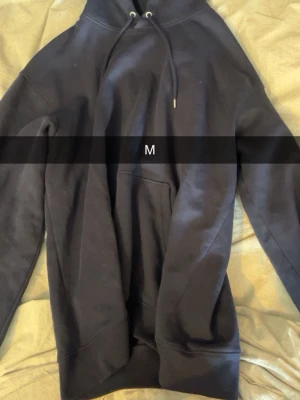 Svart hoodie med huva och ficka - En klassisk svart hoodie med huva och dragsko samt en stor magficka. Hoodien har ribbade muddar vid ärmslut och nederkant. Perfekt för en avslappnad och stilren look.