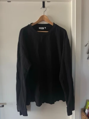 Svart sweatshirt från Weekday XL - En stilren svart tjocktröja från Weekday i storlek XL. Sweatshirten har en klassisk rund halsringning, ribbade muddar och är gjord i mjukt bomullsmaterial. Perfekt för en avslappnad och enkel look. Vid ena sidan finns en dragkedja som går att öppna typ 15cm som en designdetalj.