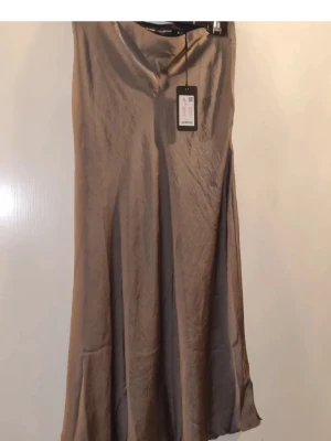 Beige långkjol i satin från Lager 157 - Stilren beige långkjol från Lager 157 i glansigt satinmaterial. Kjolen har en rak och något utsvängd passform som ger ett elegant fall. Perfekt att matcha med en enkel topp eller stickad tröja för en trendig look.