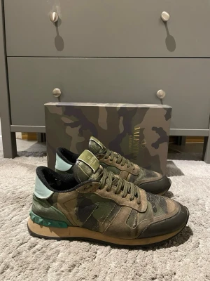Valentino camo sneakers med nitar - Snygga sneakers från Valentino i camomönstrad textil och mocka med svarta och gröna detaljer. Skorna har en unik mintgrön häl med nitar och beige sula. Perfekta för dig som vill sticka ut med exklusiv streetstyle. Storlek 42 fast passar 43 och 43,5