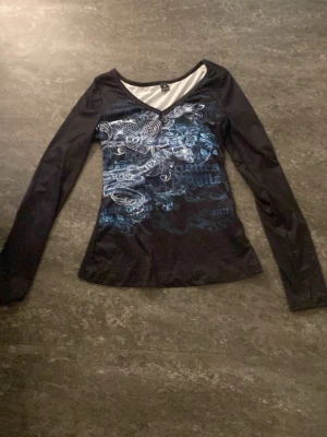 Svart långärmad topp med blått tryck - Cool svart långärmad topp med v-ringning och blått drakmönster framtill. Tunn och stretchig modell som sitter snyggt på kroppen. Perfekt för dig som gillar edgy och unika prints.