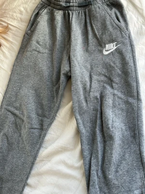 Gråa mjukisbyxor från Nike - Säljer ett par klassiska gråa mjukisbyxor från Nike med resår i midjan och sidofickor. Byxorna har Nike-loggan broderad i vitt på vänster ben och är gjorda i mjukt sweatshirtmaterial. Perfekta för chill dagar eller träning.