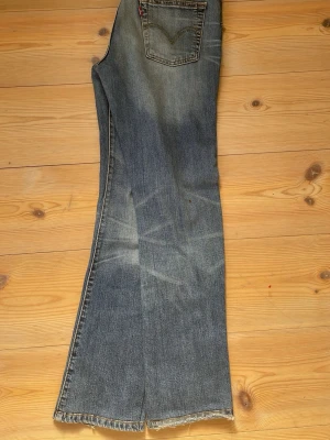 Levi's bootcut jeans med rå kant - Säljer ett par klassiska blå Levi's jeans med bootcut och rå, avklippt kant nertill. Jeansen har en snygg tvättad look, fem fickor och den ikoniska röda Levi's-taggen på bakfickan. Perfekta för en avslappnad och trendig stil.