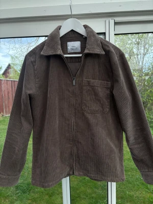 Brun overshirt i manchester från Black - Säljer en brun overshirt i manchester från Black med dragkedja framtill och en bröstficka. Overshirten har klassisk krage och raka ärmar, perfekt för lager-på-lager. Materialet är mjukt och ger en avslappnad look.