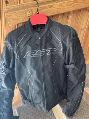 Svart motorcykeljacka från RST - Säljer en svart motorcykeljacka från RST med sportig design och diskreta grå detaljer. Jackan har lång ärm, dragkedja framtill och förstärkta partier på axlar och armbågar. Perfekt för dig som gillar snygg och funktionell stil. Använd 2-3 gånger förra säsongen 
