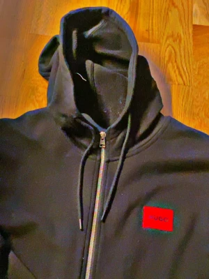 Svart hoodie med dragkedja från Hugo - Svart hoodie med hel dragkedja från Hugo. Klassisk modell med huva och dragsko, samt röd logotyp-patch på bröstet. Två fickor framtill och mjukt material som känns skönt mot huden. Perfekt för en chill och stilren look. Tagit väl hand om den cirka 2 månader använt fåtal gånger äkta såklart köptes för 2400 med dress fast mjukisen borttappade. 700kr