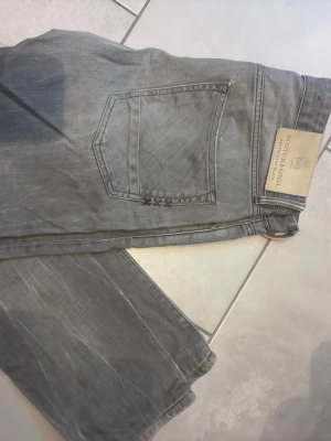 Grå jeans från Scotch & Soda - Snygga grå jeans från Scotch & Soda med klassisk femficksdesign och diskreta slitningar. Jeansen har raka ben och en patch med logga bak i midjan. Perfekta för en avslappnad och trendig look.