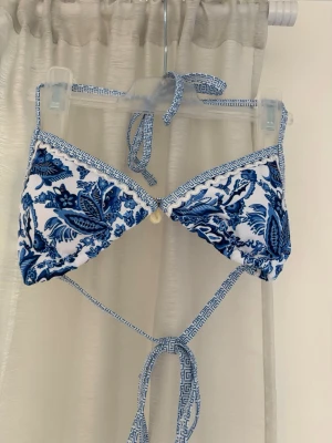  Mönstrad bikini överdel - Snygg trekantsbikini i blått och vitt med blommigt mönster och tunna knytband. Bikinin har en liten snäcka framtill och dekorativa kanter med grekiskt mönster. Perfekt för stranden eller poolen! 