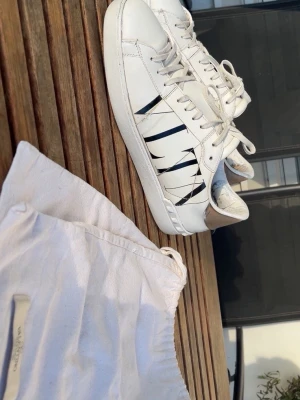 Vita Valentino Garavani sneakers - Snygga vita sneakers från Valentino Garavani med svarta detaljer på sidorna och silverfärgad häl. Klassisk låg modell med vita skosnören och gummisula. Tillverkade i läder och levereras med original dustbag.