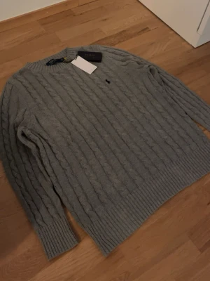 Grå kabelstickad tröja Polo Ralph Lauren - Kabelstickad grå tröja från Polo Ralph Lauren med klassisk rund hals och broderad logga på bröstet. Tröjan har ribbade muddar vid ärmslut och nederkant. Perfekt för lager-på-lager och snygg till jeans.