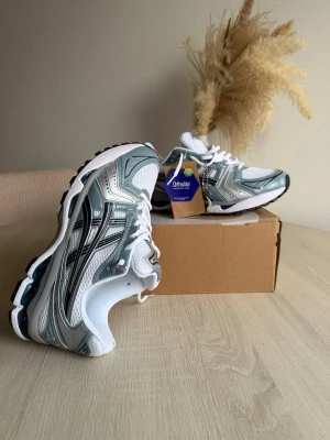 ASICS vita och silver sneakers - Snygga sneakers från ASICS i vitt och silver med svarta detaljer. Skorna har en sportig design med meshpaneler för bra ventilation och Ortholite-innersula för extra komfort. Perfekta för dig som gillar en retroinspirerad look och vill ha bekväma skor till vardags.