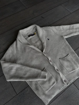 Ljusgrå kofta från Polo Ralph Lauren - Ljusgrå stickad kofta från Polo Ralph Lauren med knäppning framtill och två fickor. Klassisk sjalkrage och diskret broderad logga på bröstet. Perfekt för lager på lager och chill stil.