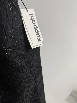 Svart ärmlös klänning med rosett Kappahl - Supersnygg svart ärmlös klänning från Kappahl i glansigt, strukturerat material. Klänningen har en djup v-ringning i ryggen och en stor dekorativ rosett bak. Tillverkad av 54% återvunnen polyester. Perfekt för dig som vill sticka ut.
