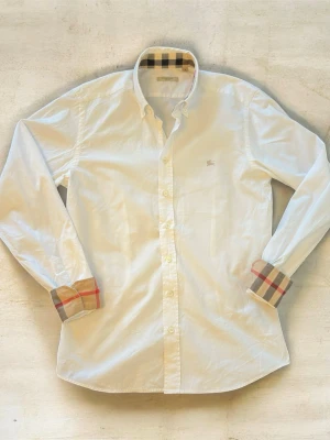 Vit skjorta från Burberry med rutiga detaljer - Klassisk vit skjorta från Burberry med ikoniska rutiga detaljer på insidan av kragen och ärmsluten. Skjortan har knappar framtill och lång ärm. Perfekt för dig som gillar stilrena plagg med en twist. Längd ca 73cm och bredd ca 47cm
