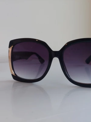 Svarta solglasögon med guld-detalj - Stilrena solglasögon med svart, fyrkantig båge och snygga gulddetaljer på sidorna. Glasen är tonade i mörkgrått till lila och ger en cool vibe till din look. Perfekta för soliga dagar och trendiga outfits.