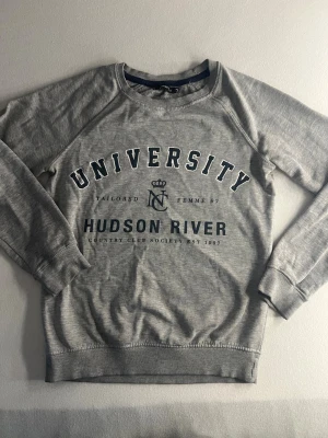Grå sweatshirt  - Snygg grå sweatshirt från Gina med tryck i mörkblått och vitt på bröstet där det står 'UNIVERSITY' och 'HUDSON RIVER'. Tröjan har rund halsringning, långa ärmar och ribbade muddar. Perfekt för en avslappnad och sportig stil.