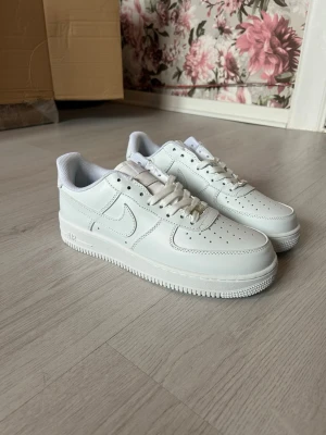 Nike Air Force 1 vita sneakers - Klassiska Nike Air Force 1 sneakers i helvitt läder med perforerad tå, vit sula och diskret Nike-logga på sidan. Skorna har lågt skaft och vita skosnören. Perfekta för en clean och stilren look.