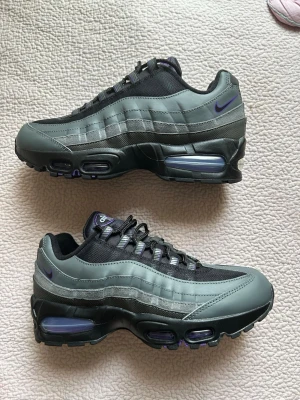 Nike Air Max 95 grå/svarta sneakers - Nike Air Max 95 sneakers i grått och svart med lila detaljer. Ovandelen är i mesh och syntet med vågiga paneler, synlig Air-enhet i sulan och broderad Swoosh på sidan. Klassisk chunky siluett och snörning framtill. Perfekta för dig som gillar streetwear.