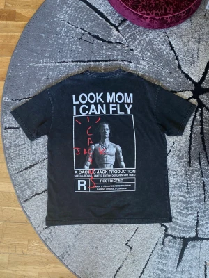 Svart Cactus Jack T-shirt Look Mom I Can Fly - Svart T-shirt från Cactus Jack med stort tryck på ryggen: 'LOOK MOM I CAN FLY' och en bild av Travis Scott. Text och grafiska detaljer i vitt och rött. Stonewashed. Perfekt statement-plagg för dig som gillar streetwear. 