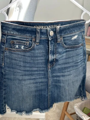 American Eagle jeanskjol - Säljer en jeanskjol från American Eagle, nytt skick med dekorativa fickor fram och bak. Från toppen till botten av kjolen är den totalt 36cm i längd. Stretchig och perfekt för sommaren