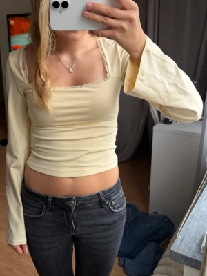 Gul långärmad topp med rosett - Söt ljusgul långärmad topp med fyrkantig ringning och spetsdetaljer. Toppen har en öppen rygg med knytrosett bak. Super skönt material från Gina Young 