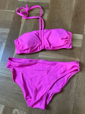 Neonrosa bandeau bikini - Neonrosa bikini med bandeau-topp och avtagbara axelband. Klassiska bikini trosor som är helt oanvända. Materialet är stretchigt och snabbtorkande. Storlek S på både topp och trosor. 