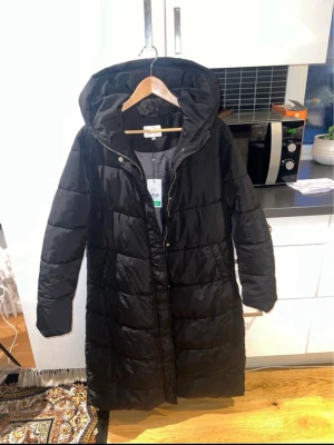 Svart lång dunjacka från Esprit - Snygg svart lång dunjacka från Esprit med huva och quiltad design. Jackan har dragkedja och tryckknappar framtill, samt två fickor på sidorna. Perfekt för kalla dagar och enkel att styla till olika outfits.
