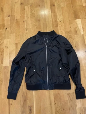 Svart bomberjacka Jack & Jones - Snygg svart bomberjacka från Jack & Jones Core med silvrig dragkedja och två fickor med tryckknappar. Jackan har ribbade muddar vid ärmslut, krage och nederkant. Tillverkad i glansigt syntetmaterial som ger en modern look.obs flärpen till dragkedjan saknas annars fint skick