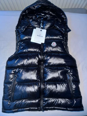 Svart dunväst från Moncler med huva - Snygg svart dunväst från Moncler med glansig finish, huva och två dragkedjefickor. Västen har Moncler-logga på bröstet och stängs med dragkedja. Perfekt för lager-på-lager och streetstyle. Aldrig använd helt ny (har bara en liten repa på sista bilden) 