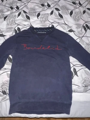 Mörkblå sweatshirt från Bondelid - Mörkblå sweatshirt från Bondelid med röd broderad logga över bröstet. Tröjan har en avslappnad passform, ribbade muddar och en unik V-detalj vid halsringningen. Perfekt för en casual och stilren look.