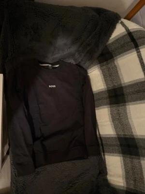 Svart sweatshirt från BOSS - Svart sweatshirt från BOSS med rund halsringning och vit broderad logotyp på bröstet. Tröjan har en klassisk passform och är tillverkad i mjukt bomullsmaterial. Perfekt för en stilren och avslappnad look.