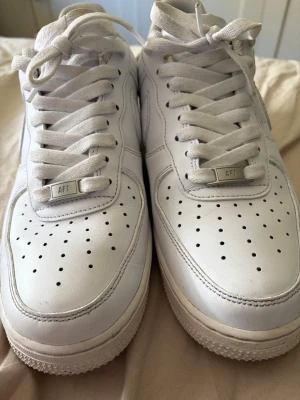 Nike Air Force 1 vita sneakers - Klassiska Nike Air Force 1 sneakers i helvitt läder med perforerad tå, vita skosnören och diskret Nike-logga bak. Snygg och tidlös modell med låg profil och robust sula. Perfekta för en clean och stilren look.