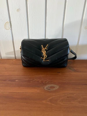 Svart YSL handväska i läder - Säljer en svart handväska från Saint Laurent med klassisk YSL-logga i guld framtill. Väskan är tillverkad i quiltat läder och har en axelrem i matchande svart med gulddetaljer. Perfekt storlek för det viktigaste och riktigt snygg design.