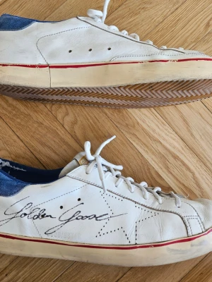 Golden Goose Super-star vita sneakers, st 43 - Golden Goose sneakers i vitt läder med blå häl och röd rand längs sulan. Klassisk låg modell med snörning, stjärndetalj på sidan och märkets logga i svart. Skorna är välanvända med synlig slitage på hälparti (invändigt) och baktill på sulan. Nypris 6000kr