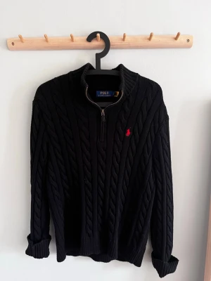 Svart kabelstickad tröja Polo Ralph Lauren - Svart kabelstickad tröja från Polo Ralph Lauren med halv dragkedja och röd broderad logga på bröstet. Tröjan har hög krage, långa ärmar och ribbade muddar. Perfekt för lager-på-lager och klassisk stil.