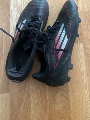 Adidas F50 svarta fotbollsskor - Säljer ett par svarta Adidas F50 fotbollsskor med röda och vita detaljer samt klassiska tre ränder på sidan. Skorna har snörning och dobbar för spel på gräs. Snygg, sportig design i syntetmaterial som ger bra grepp och känsla på planen. Pris kan diskuteras de är inte använda köpte de och de är för små sen har glömt att lämna in dem!